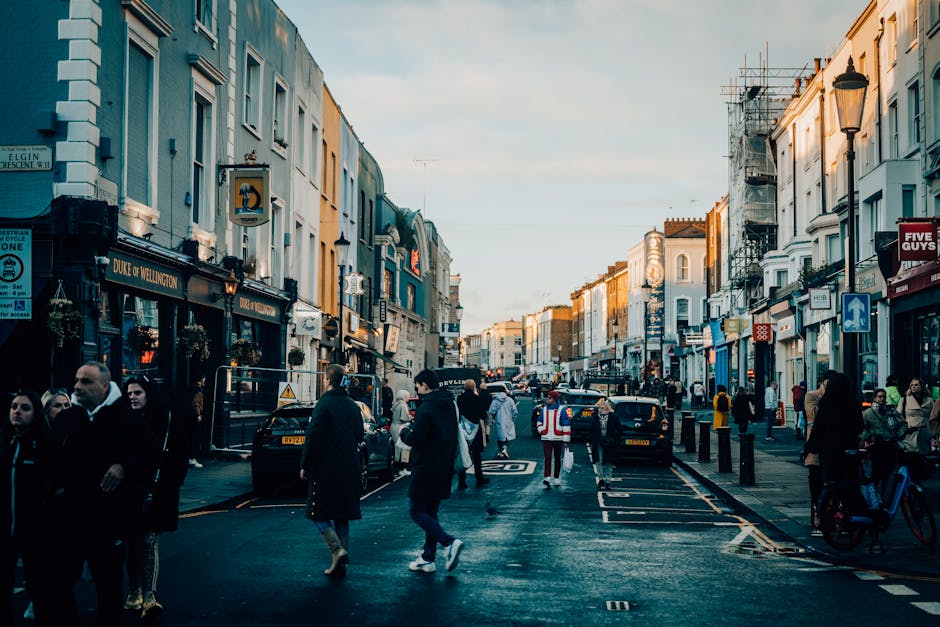 A busy street scene in Notting Hill during daylight, featuring a variety of retail shops with black and white signage, pedestrians walking on the wet pavement, some in coats and scarves, and cars park