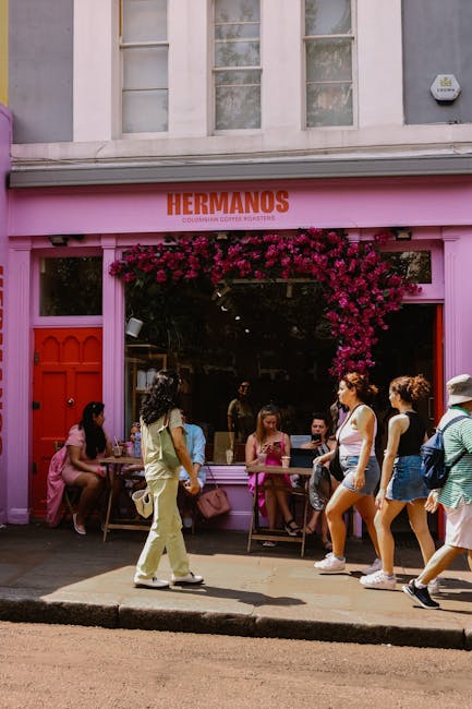 A vibrant storefront of Hermanos, a coffee shop painted in bright pink with a prominent sign above reading 'Hermanos.' The shop is decorated with a lush, cascading arrangement of deep pink and burgund