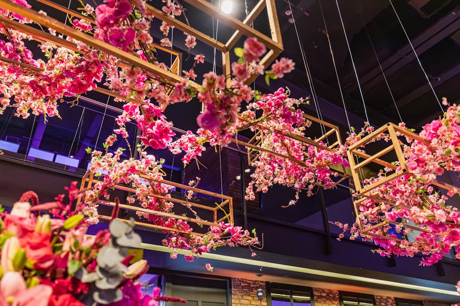 An indoor installation of vibrant pink cherry blossom branches arranged within geometric wooden frames hanging from the ceiling. The flowers display various shades of pink with some white accents, fea