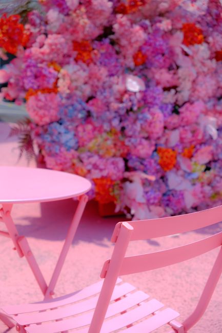 A close-up of a floral arrangement consisting of densely packed pink, purple, and orange flowers, creating a vibrant, textured backdrop. In the foreground, there are two pastel pink chairs with slatte