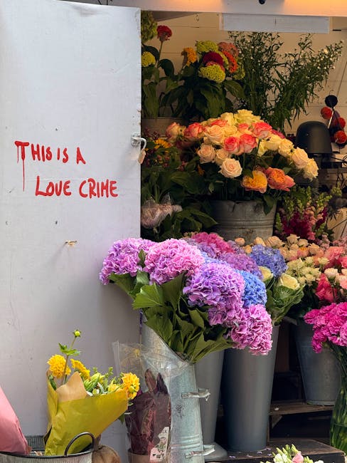 A vibrant display of fresh flowers at a florist shop, featuring a bouquet of pink and purple hydrangeas in a clear plastic wrapping, placed in a tall, silver vase. Behind, arrangements of roses, chrys