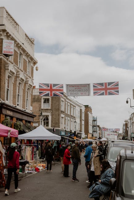 A bustling outdoor scene at Portobello Market with various flower stalls and vendors. Brightly colored floral arrangements including roses, peonies, and lilies are displayed, wrapped in elegant paper