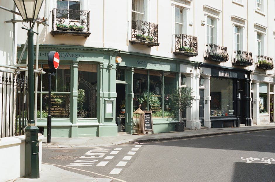 An exterior view of a floral boutique located on a street corner in Notting Hill, showcasing a soft pastel green facade with large display windows filled with fresh floral arrangements. The shop featu