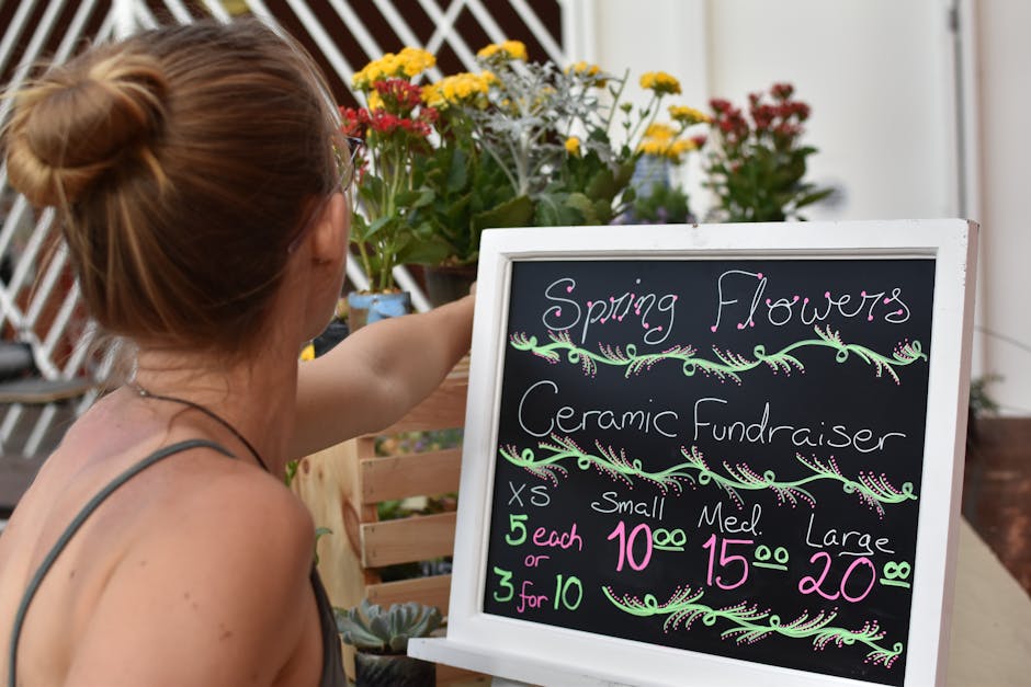 A woman with reddish-brown hair styled in a bun, wearing a gray tank top, is pointing at a blackboard sign advertising spring flowers and ceramic fundraisers outside a flower shop. The sign displays h