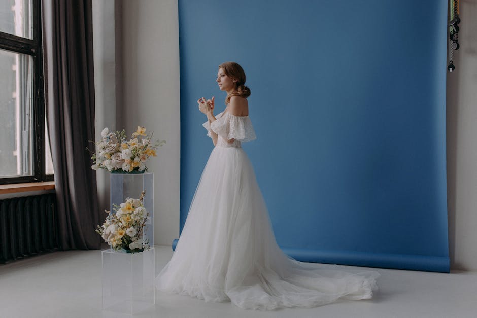 A woman in a white wedding dress with off-the-shoulder lace sleeves stands indoors against a blue backdrop, holding a smartphone. To her side, there are two arrangements of fresh floral bouquets featu