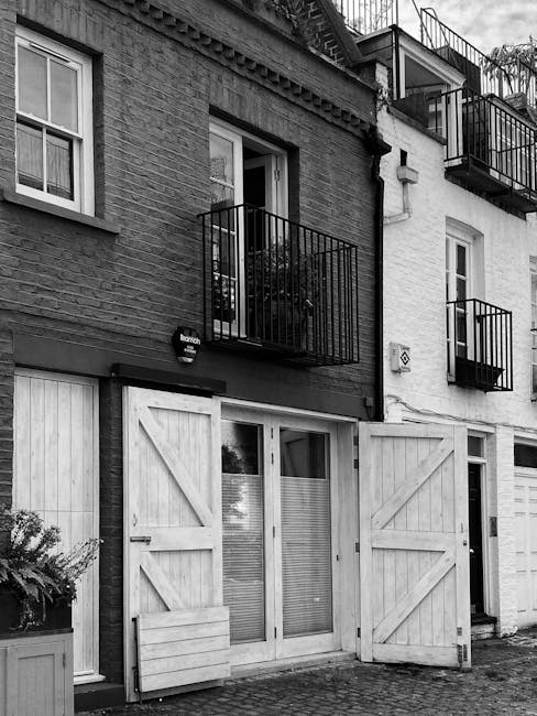 The image depicts the facade of a multi-story residential building in black and white, featuring a ground-level glass door with wooden barn-style shutters partially opened, revealing a glimpse of the 